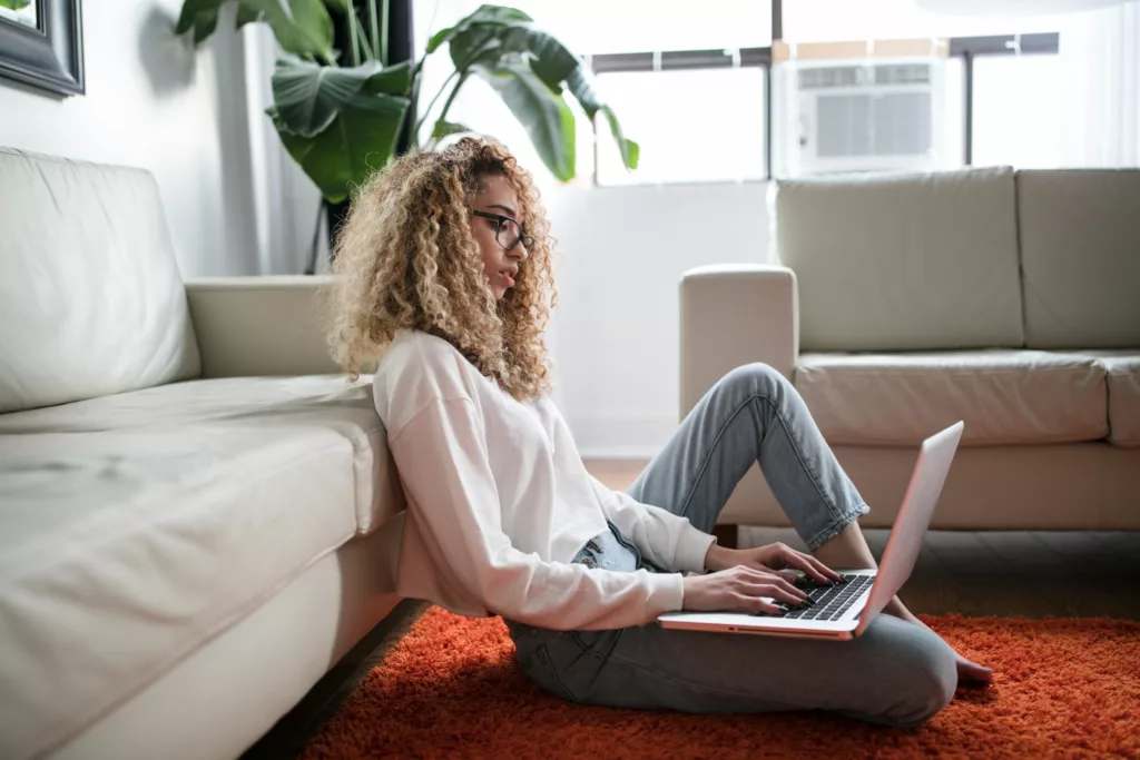 Ung kvinne sitter på gulvet inntil en sofa med en PC i fanget